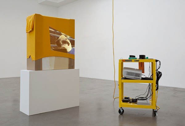 Rachel Harrison, AA, 2010. Wood, bubble wrap, cardboard, acrylic, tennis shirt, A/V cart, DVD player, speakers, projector, extension cord, five hair rollers, pack of gum, ear plugs, and American Apparel video, color, sound, 18 minutes (2009). Courtesy of the artist, Greene Naftali, New York and Regen Projects, Los Angeles. Photography by Brian Forrest.
